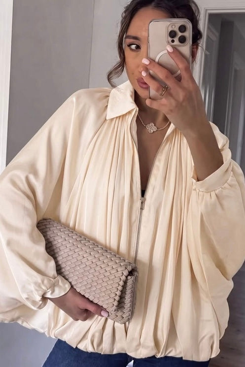 A woman takes a mirror selfie wearing a cream-colored, voluminous pleated blouse with a front zipper and holding a woven beige clutch.