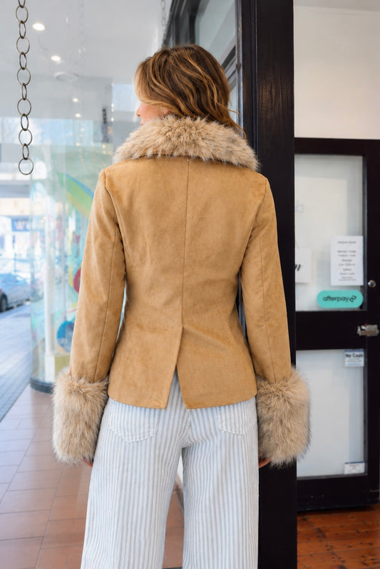 A woman viewed from behind wearing a tan suede jacket with a light brown fur collar and cuffs, paired with white and gray pinstriped trousers, standing in front of a storefront.