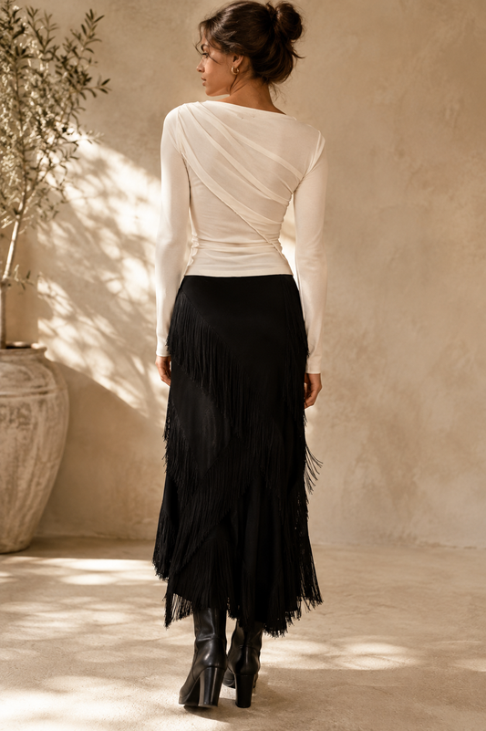 A woman shown from behind wearing a cream off-the-shoulder long-sleeve top and a black fringe midi skirt, paired with black boots, in a sunlit rustic room with a textured beige wall.
