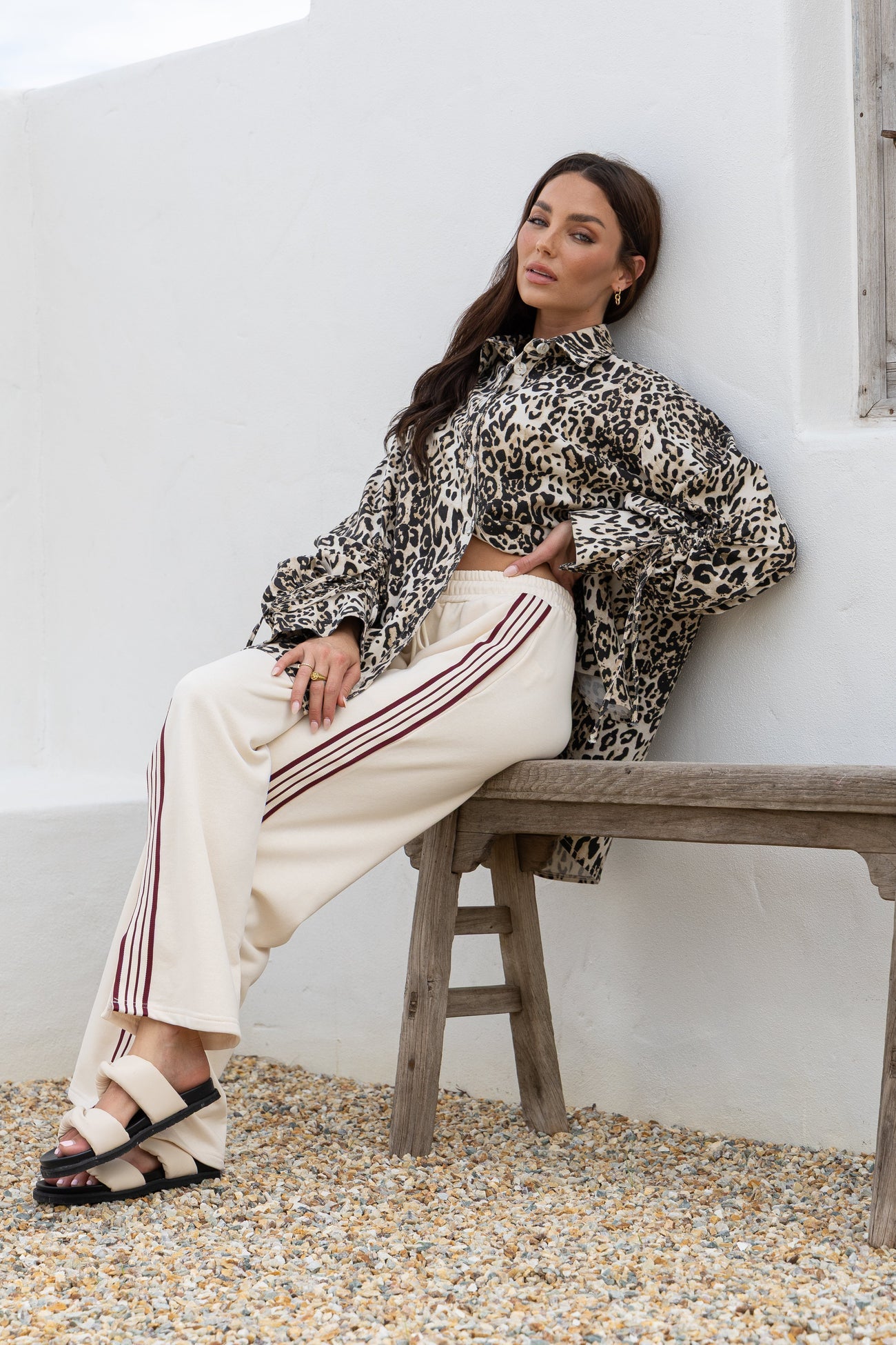 Woman sitting on a wooden bench wearing a leopard print top and cream pants with red stripes.