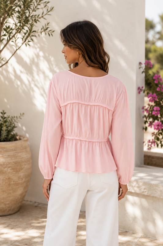 A woman wearing a light pink long-sleeve blouse with a gathered peplum-style back, paired with white pants, photographed outdoors against a sunlit white wall with potted plants.
