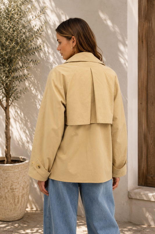 A woman wearing a beige trench coat with a unique double-breasted back detail and light blue jeans, photographed from behind in an outdoor setting with a potted tree and white stucco wall.