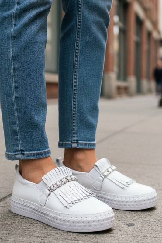 White slip-on fringed loafers with rhinestone straps, worn with blue jeans on a city sidewalk.