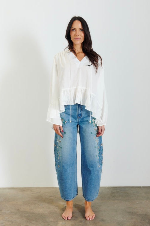 A woman stands barefoot against a plain white wall wearing a loose white long-sleeve blouse and light blue cropped jeans with floral embroidery on the front.