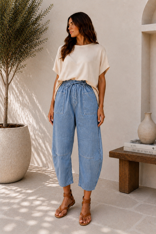 A woman stands in a sunlit indoor-outdoor setting wearing a cream short-sleeve top, light blue high-waisted cropped wide-leg denim pants, and brown strappy sandals.