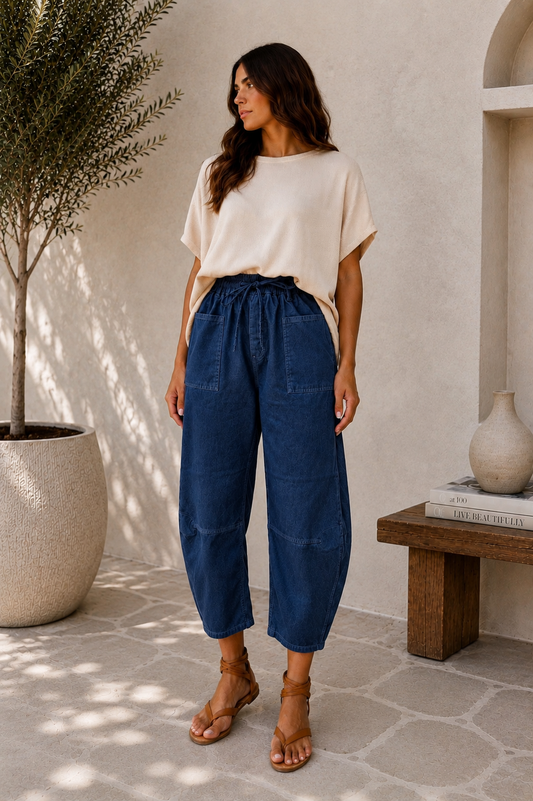 A woman stands in a sunlit indoor-outdoor space wearing loose dark blue wide-leg cropped pants with a black drawstring waist, a cream short-sleeve top, and tan sandals. A potted tree, textured wall, bench, and vase are visible in the background.