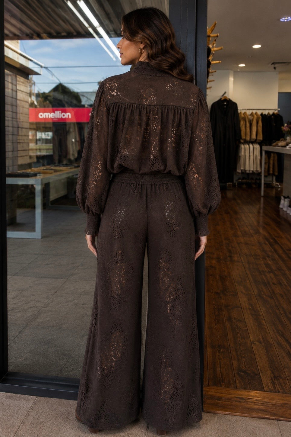 A woman stands in a clothing store wearing a dark brown lace jumpsuit with a high neck, long sleeves, and wide legs. The back of the jumpsuit features a ruffled yoke and subtle floral lace patterns.