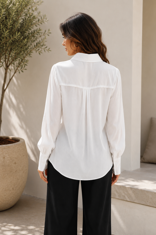 A woman wearing a white long-sleeve blouse with a high collar and gathered cuffs, paired with black wide-leg pants, shown from the back in a sunlit indoor-outdoor setting.