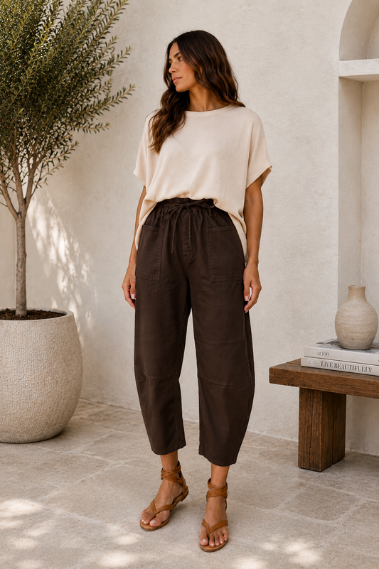 A woman stands in a sunlit indoor-outdoor space wearing loose beige short-sleeve top and dark brown high-waisted cropped pants with a drawstring waist, paired with tan sandals.