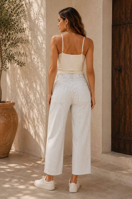 A woman viewed from behind wearing a cream camisole and white pinstriped wide-leg cropped pants, standing in a sunlit outdoor setting with a stucco wall, wooden door, and potted olive tree.