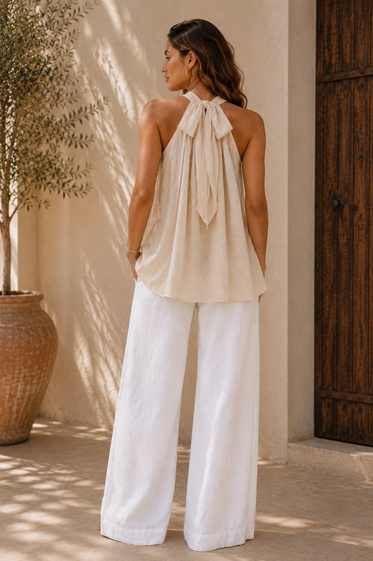 A woman stands outdoors wearing a sleeveless halter-style top with a large bow at the back and wide-leg white pants, photographed against a sunlit beige wall and wooden door.