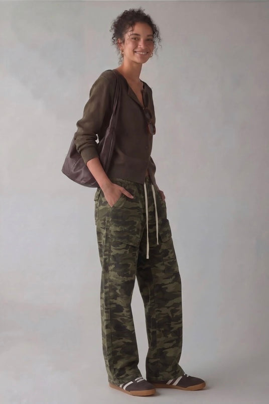 A woman stands against a light gray studio background wearing a dark olive-brown long-sleeve top, loose camouflage drawstring pants in green and brown tones, and brown slip-on shoes. She is carrying a dark shoulder bag.