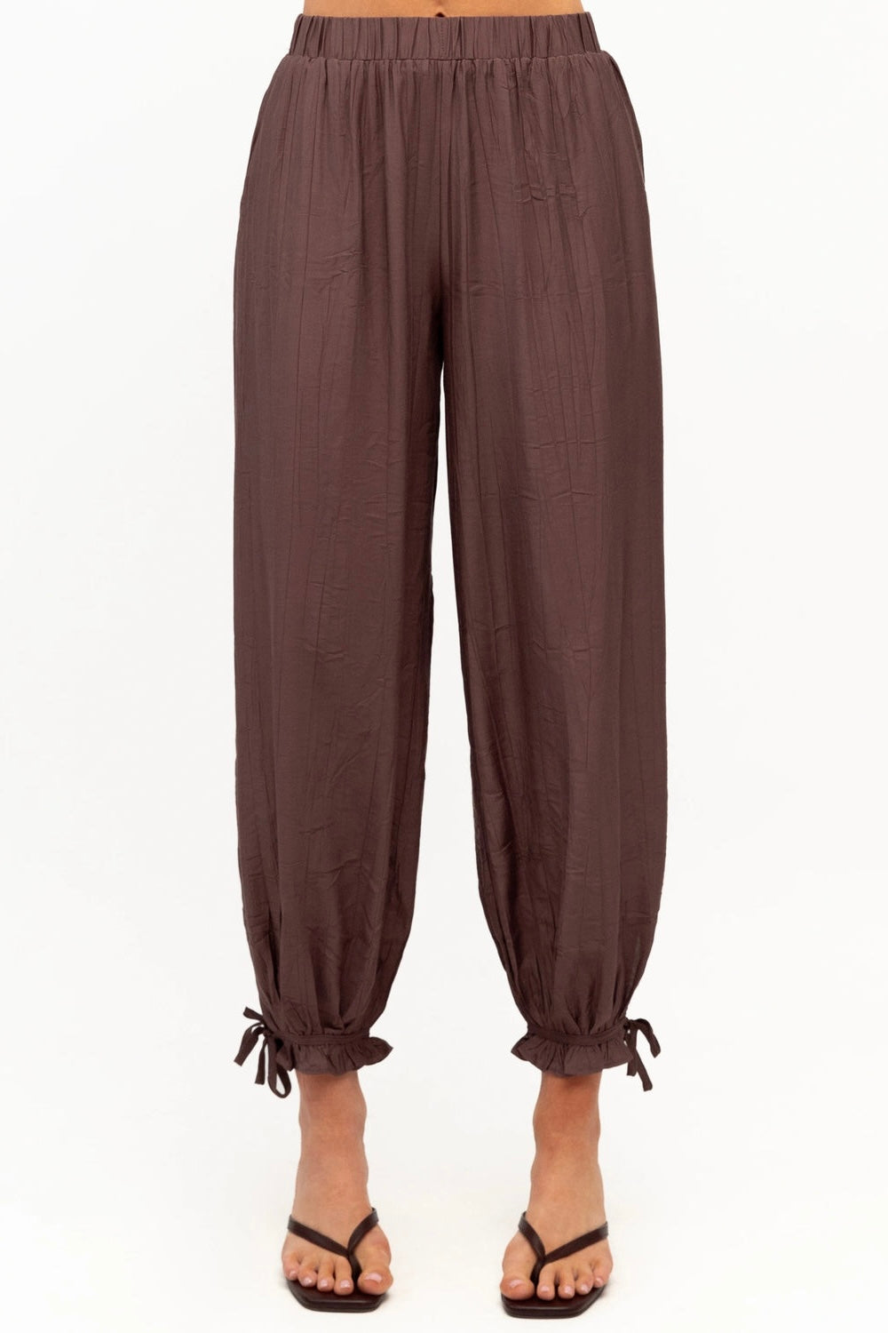 A pair of dark brown, loose-fitting harem-style pants with a gathered elastic waist and drawstring ankle cuffs, shown on a model wearing black flip-flop sandals against a plain white background.