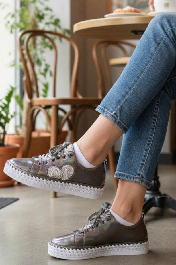 A person wearing metallic taupe-brown lace-up platform sneakers with a heart-shaped rhinestone embellishment on the side, paired with blue jeans in a cafe setting.
