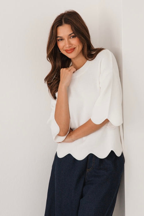 Woman wearing a white knit with scalloped edges and dark pants against a light background