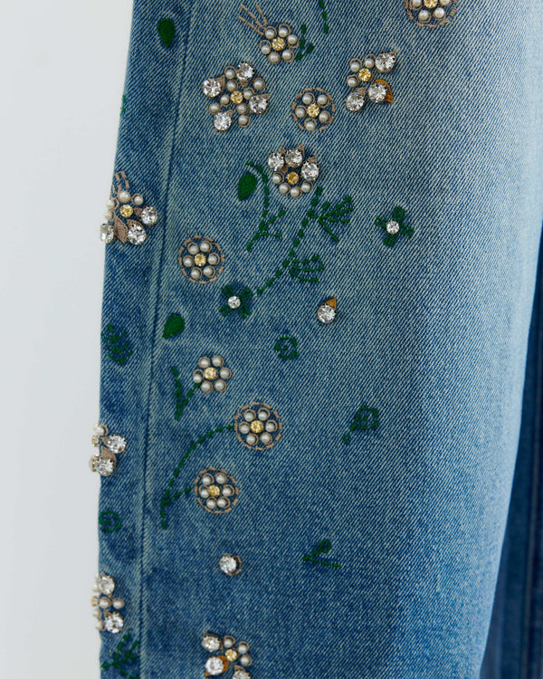Close-up of blue denim fabric with embroidered floral details and rhinestone embellishments, including green leaves and white, yellow, and silver flower motifs.