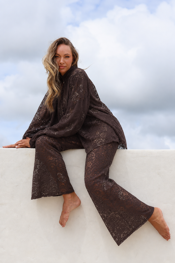 Woman wearing a dark brown lace outfit sitting on a white ledge with a cloudy sky background
