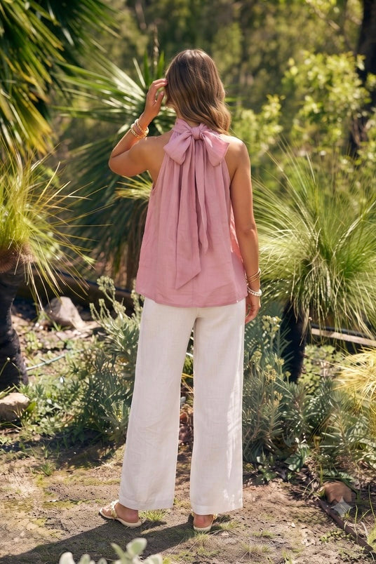 A woman stands outdoors in a garden wearing a sleeveless dusty pink top with a large bow at the back of the neck and white wide-leg pants.