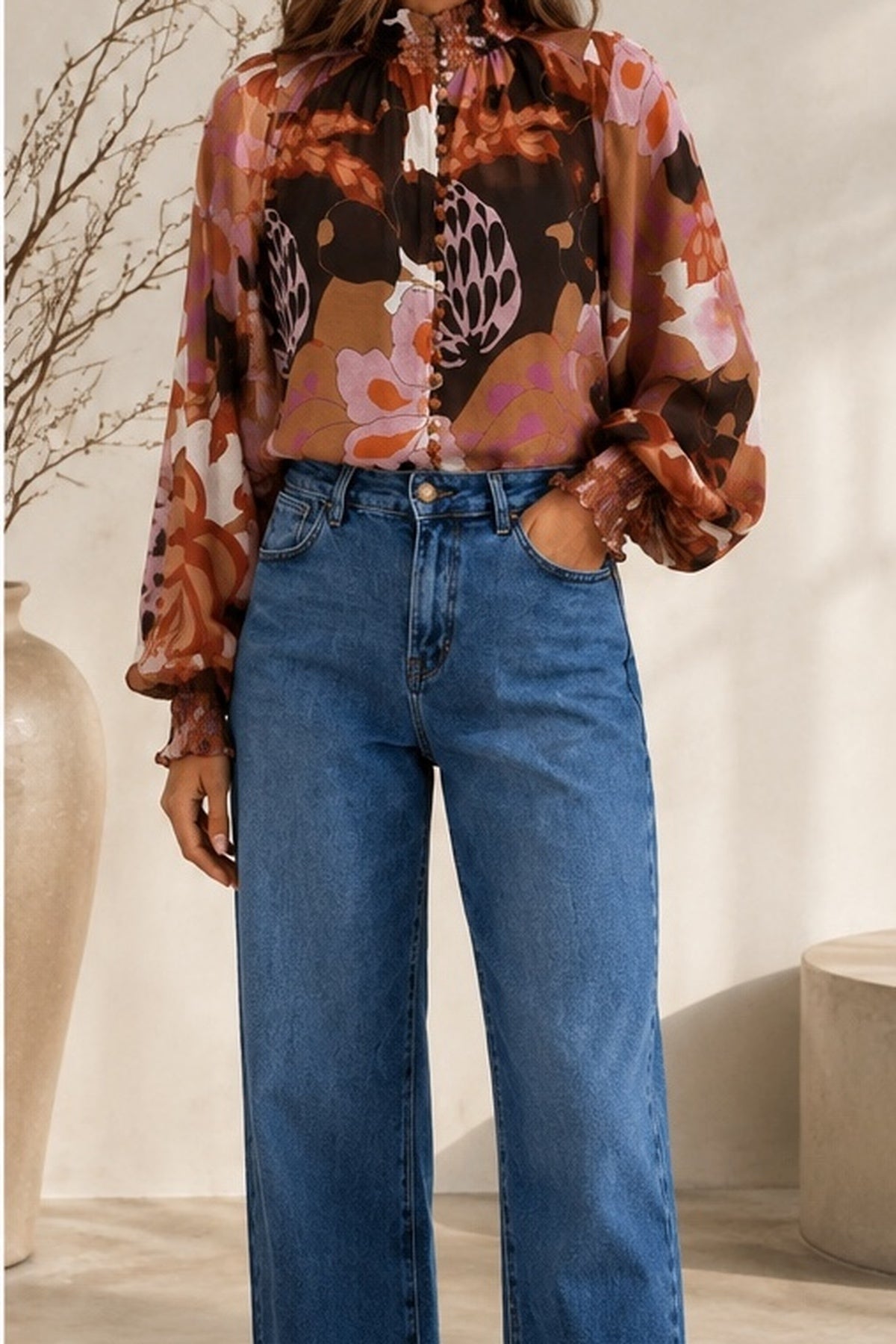 A woman wearing a sheer long-sleeve blouse with a brown, pink, and black abstract floral pattern, tucked into high-waisted blue wide-leg jeans, standing in a brightly lit indoor setting.