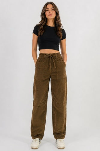 A woman modeling a pair of high-waisted, wide-leg brown corduroy pants with a drawstring waist, paired with a black crop top and white sneakers, against a plain light gray background.