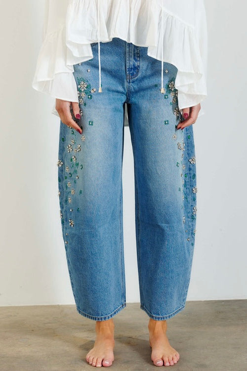 A person wearing light blue wide-leg cropped jeans with floral bead embellishments down the sides, paired with a white ruffled blouse and bare feet, standing against a plain light wall.