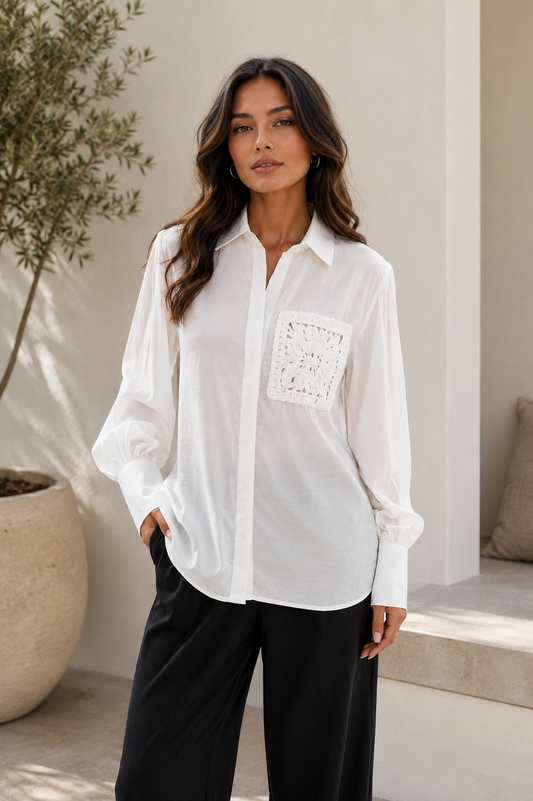 A woman wearing a white long-sleeve button-up shirt with a square lace-up chest pocket, paired with black wide-leg pants, standing in a sunlit modern outdoor setting.