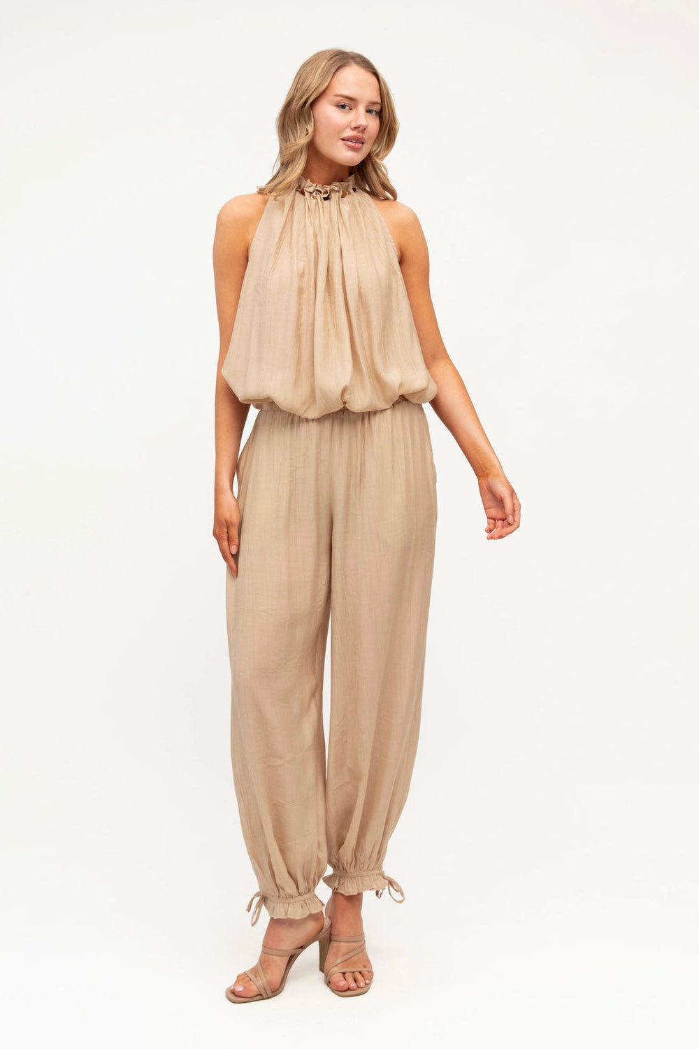 A woman modeling a sleeveless beige jumpsuit with a gathered halter-style top and loose, wide-leg pants with tie details at the ankles, photographed against a plain white studio background.