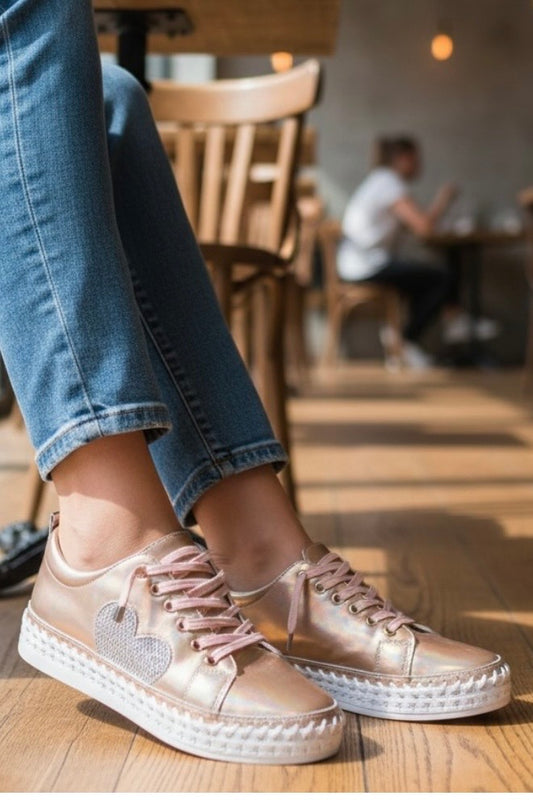A person wearing metallic rose gold low-top sneakers with a heart-shaped rhinestone on the side, paired with blue jeans in a warm, sunlit cafe setting.