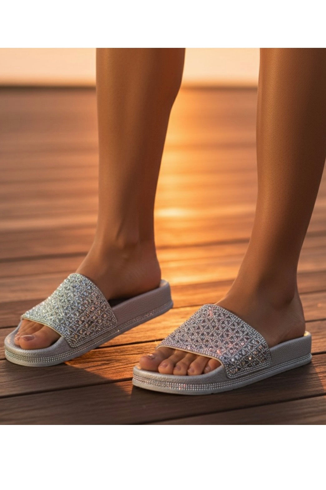 A pair of women's slide sandals with sparkling silver rhinestone-embellished straps and white soles, shown being worn on feet on a wooden deck at sunset.