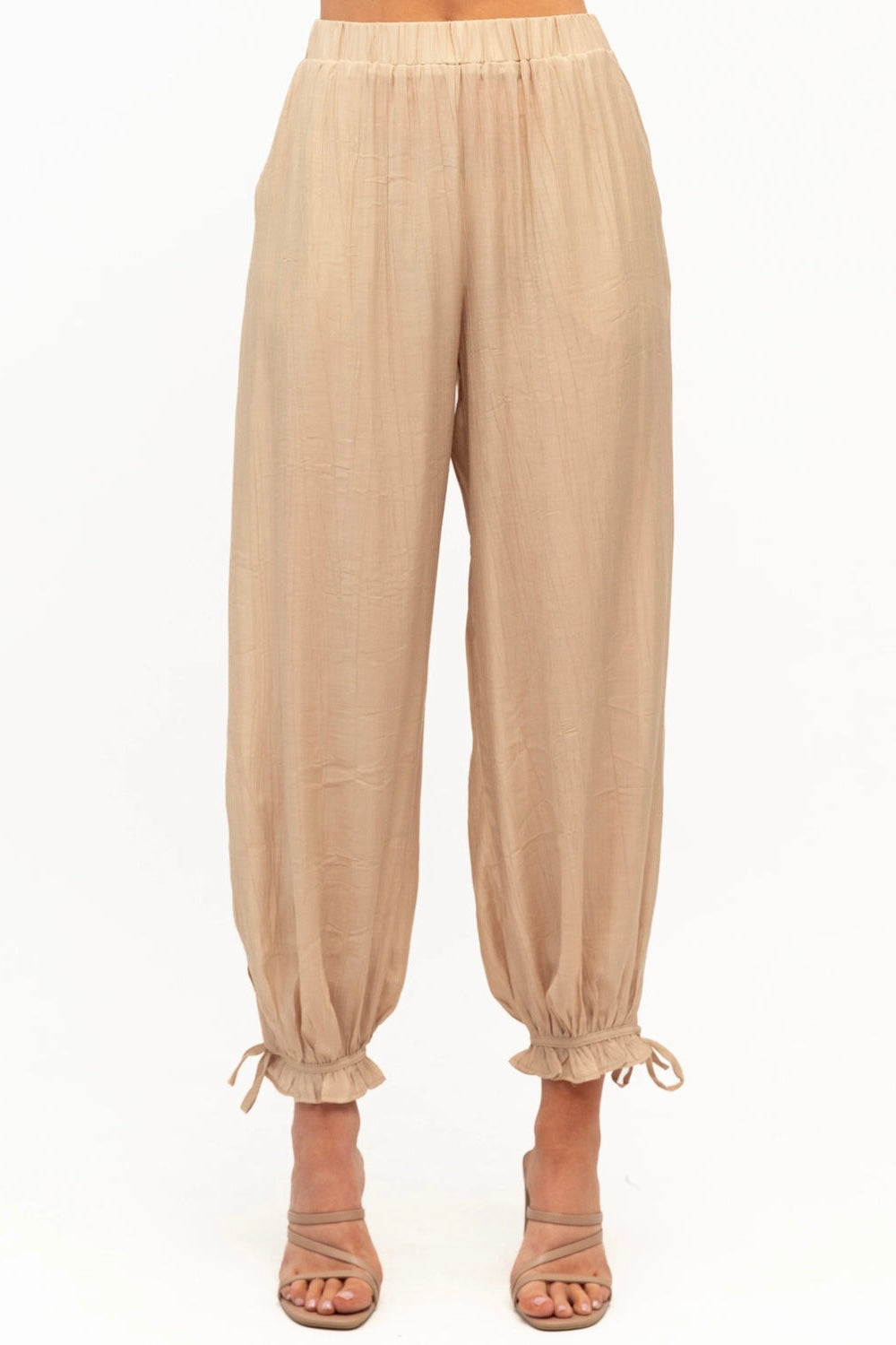 Beige high-waisted, loose-fitting pants with a drawstring waist and ruffled ankle cuffs, shown on a model wearing beige strappy heels against a white background.
