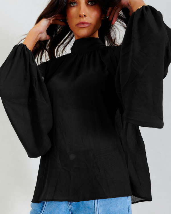 A woman wearing a black long-sleeve blouse with dramatic balloon sleeves and a high gathered neckline, paired with light blue jeans, against a plain light gray background.