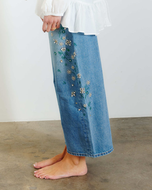 A person wearing a cropped blue denim skirt with floral embroidery down the side, paired with a white ruffled top and bare feet on a concrete floor against a white wall.