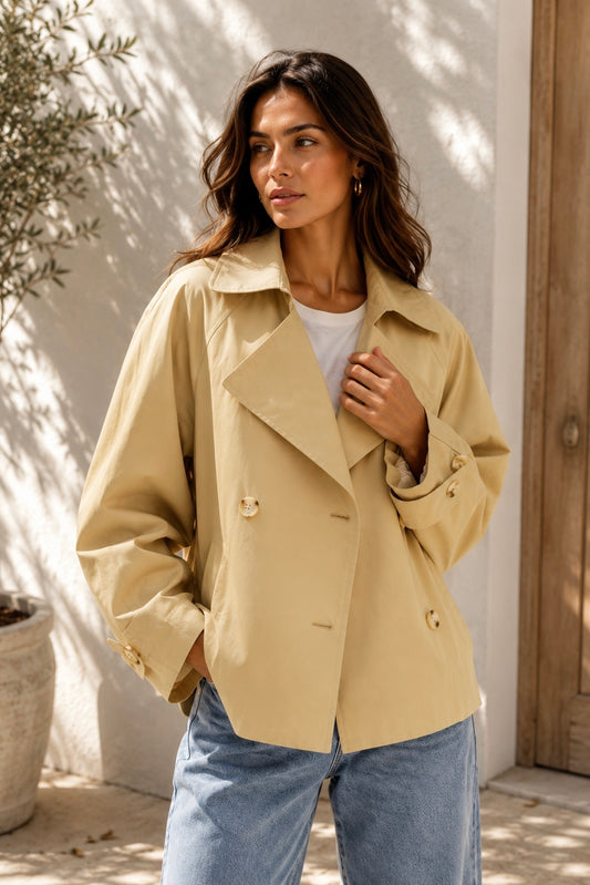 A woman wearing a light beige double-breasted trench coat over a white t-shirt and blue jeans, standing outdoors against a sunlit stucco wall.