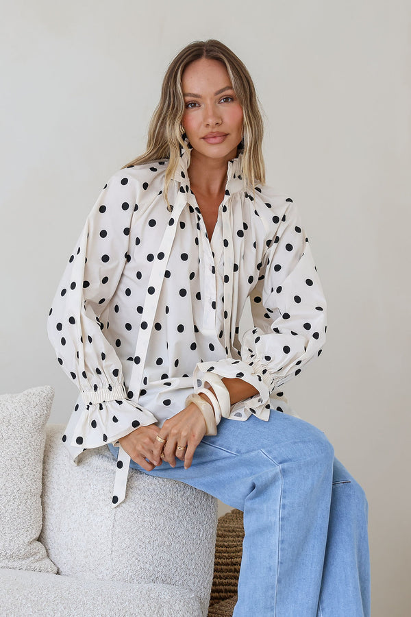 Woman wearing a polka dot blouse and light blue jeans sitting on a couch.
