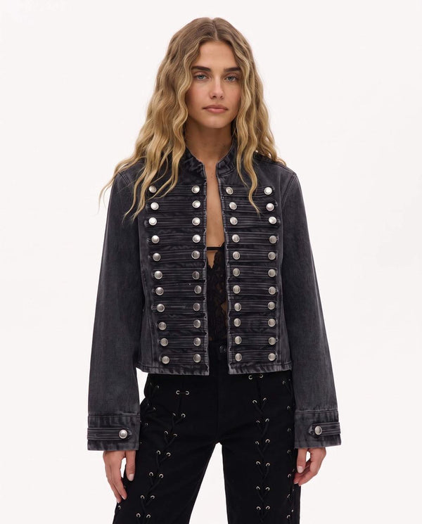 A woman modeling a cropped black washed denim jacket with double-breasted silver military-style buttons, paired with black studded pants, against a plain white studio background.