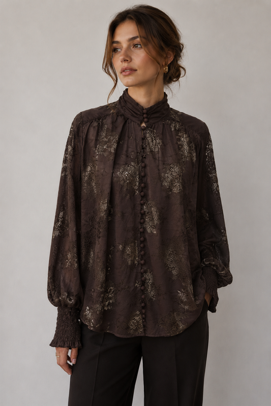 A woman wearing a dark brown long-sleeve blouse with a subtle tonal floral pattern, high gathered collar, and button front, paired with black pants, against a neutral gray background.