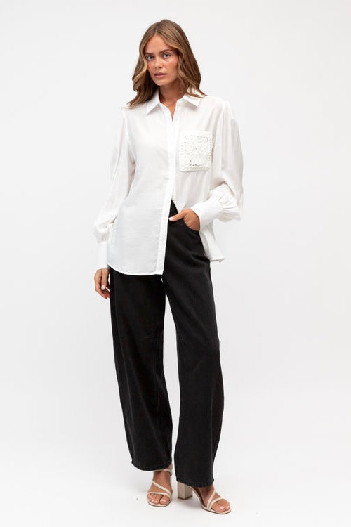 A woman modeling a white long-sleeve button-up shirt with a decorative embroidered chest pocket, paired with black wide-leg pants and beige heeled sandals, against a plain white studio background.