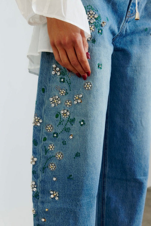 Close-up of a person wearing blue jeans with floral bead and sequin embellishments on the left leg, paired with a white blouse.