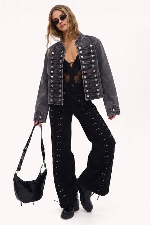 A model wearing a washed black denim jacket with silver button details, black studded wide-leg pants, and black boots, holding a black shoulder bag against a plain white background.