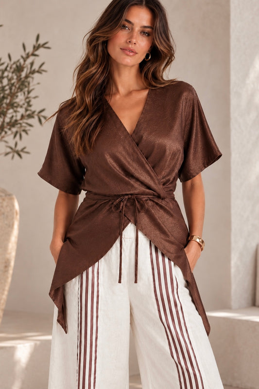 A woman wearing a brown satin-like short-sleeve wrap top with a tie waist, paired with white pants featuring vertical brown stripes, in a softly lit indoor setting.