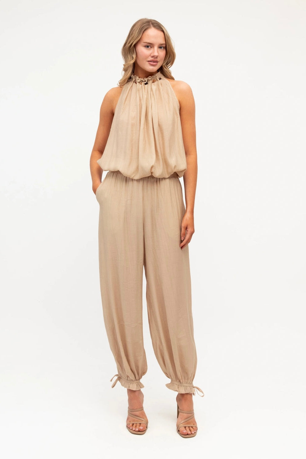 A woman modeling a sleeveless beige halter jumpsuit with a gathered, ruffled bodice and loose, drawstring-hem pants, paired with strappy heeled sandals against a plain white studio background.