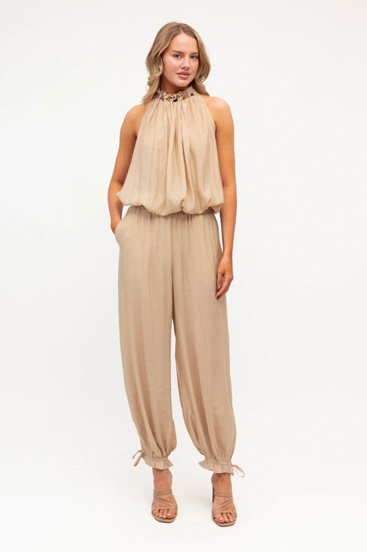 A woman modeling a sleeveless beige halter jumpsuit with a gathered, ruffled bodice and loose, drawstring-hem pants, paired with strappy heeled sandals against a plain white studio background.
