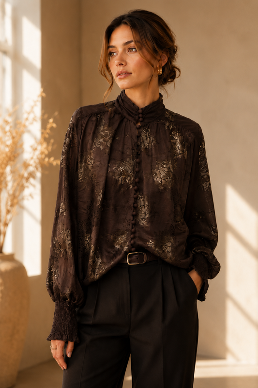 A woman wearing a dark brown sheer long-sleeve blouse with a subtle gold floral pattern, styled with black trousers in a warm, sunlit interior setting.