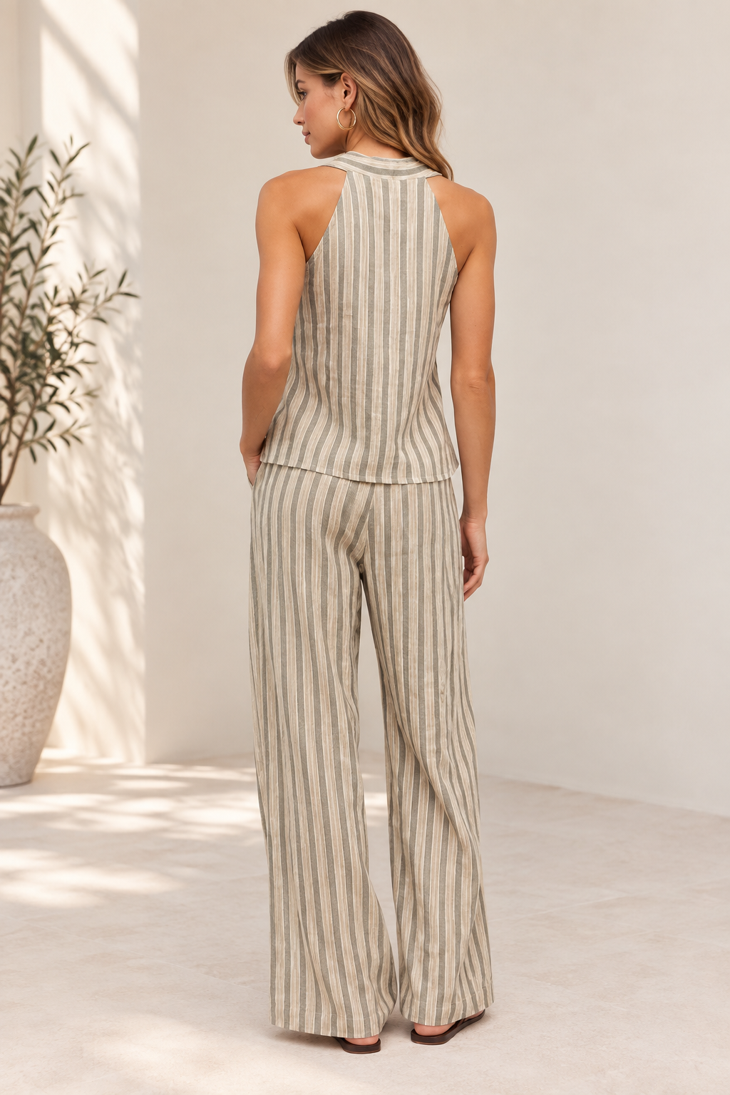 A woman stands with her back to the camera wearing a sleeveless beige and cream vertical-striped halter top and matching wide-leg pants in a bright, minimalist room.