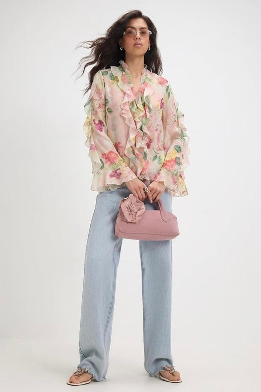A woman modeling a light pink floral long-sleeve blouse with ruffled details, paired with light blue wide-leg jeans, a dusty pink handbag, and brown loafers, against a plain white studio background.