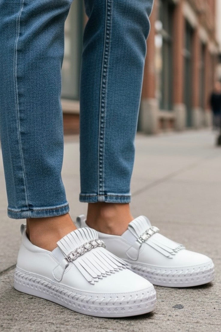 White slip-on fringed loafers with rhinestone straps, worn with blue jeans on a city sidewalk.