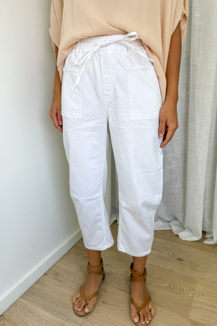 White cropped pants with a drawstring waist and large patch pockets, shown on a model wearing a beige top and brown sandals in an indoor setting.