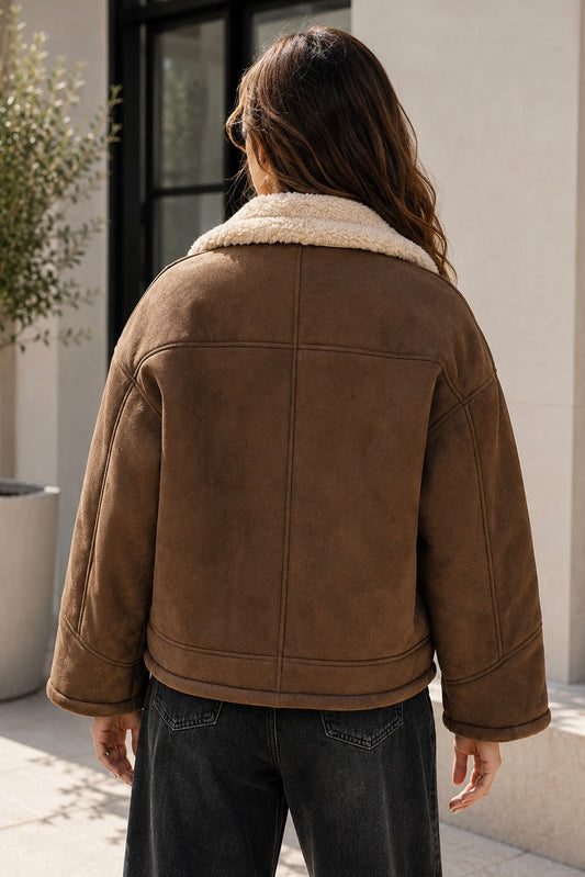 A person viewed from behind wearing a brown suede-like jacket with a cream sherpa collar, paired with dark jeans, standing outdoors against a light-colored building wall.