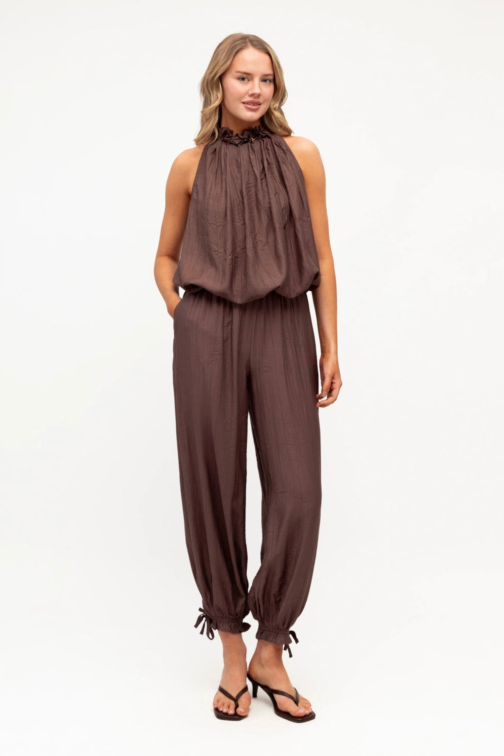 A woman modeling a sleeveless high-neck jumpsuit in a dark brown color, paired with black thong sandals, against a plain white studio background.