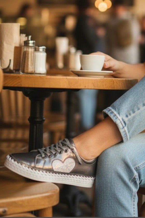 A person sitting at a wooden cafe table wearing metallic silver low-top sneakers with a glittery heart on the side. The person is wearing light blue jeans, and a white coffee cup sits on the table in front of them.